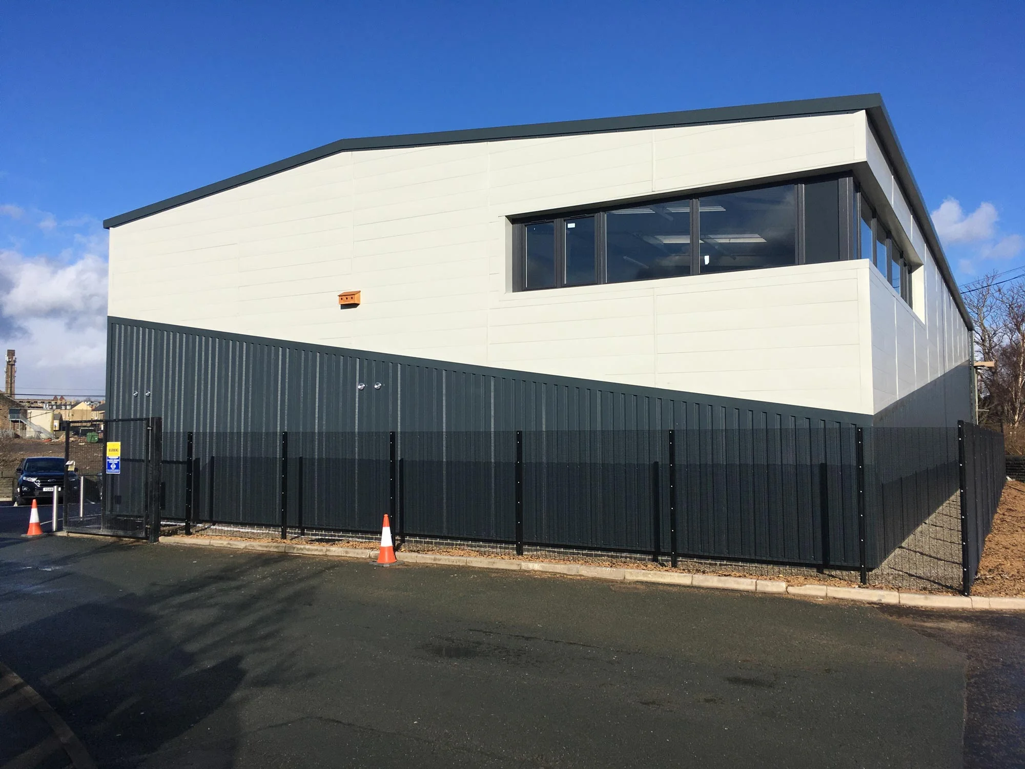 A modern industrial building with large windows, grey metal cladding, and a black metal fence—expertly designed by structural and civil engineers—stands on a paved car park under a clear blue sky.