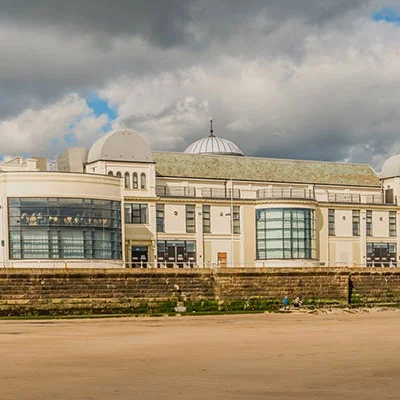 A large, white, domed building with arched windows and a stone wall in front, set against a cloudy sky and sandy ground, showcases impressive structural design admired by engineers and surveying professionals alike.