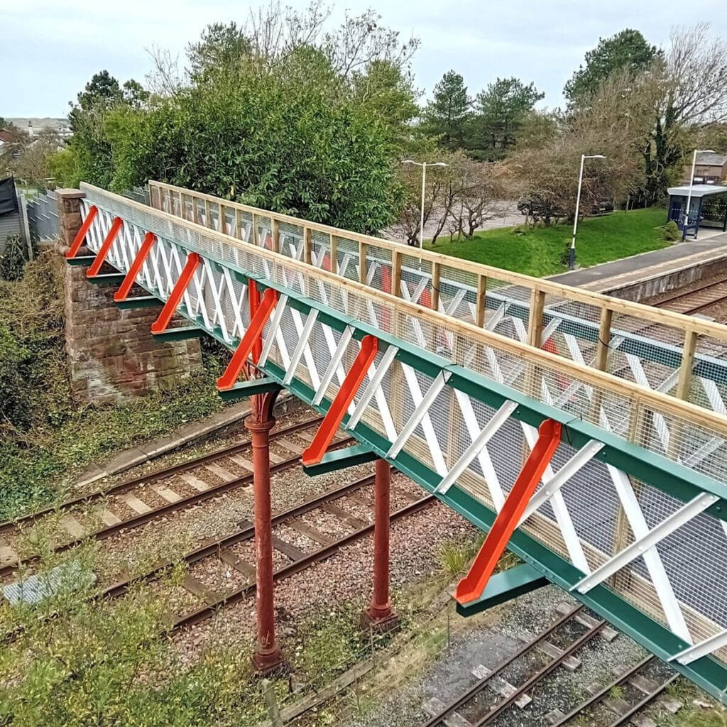 A pedestrian bridge with a metal lattice structure and wooden railing spans over railway tracks at a small, leafy railway station. Ravensglass Footbridge