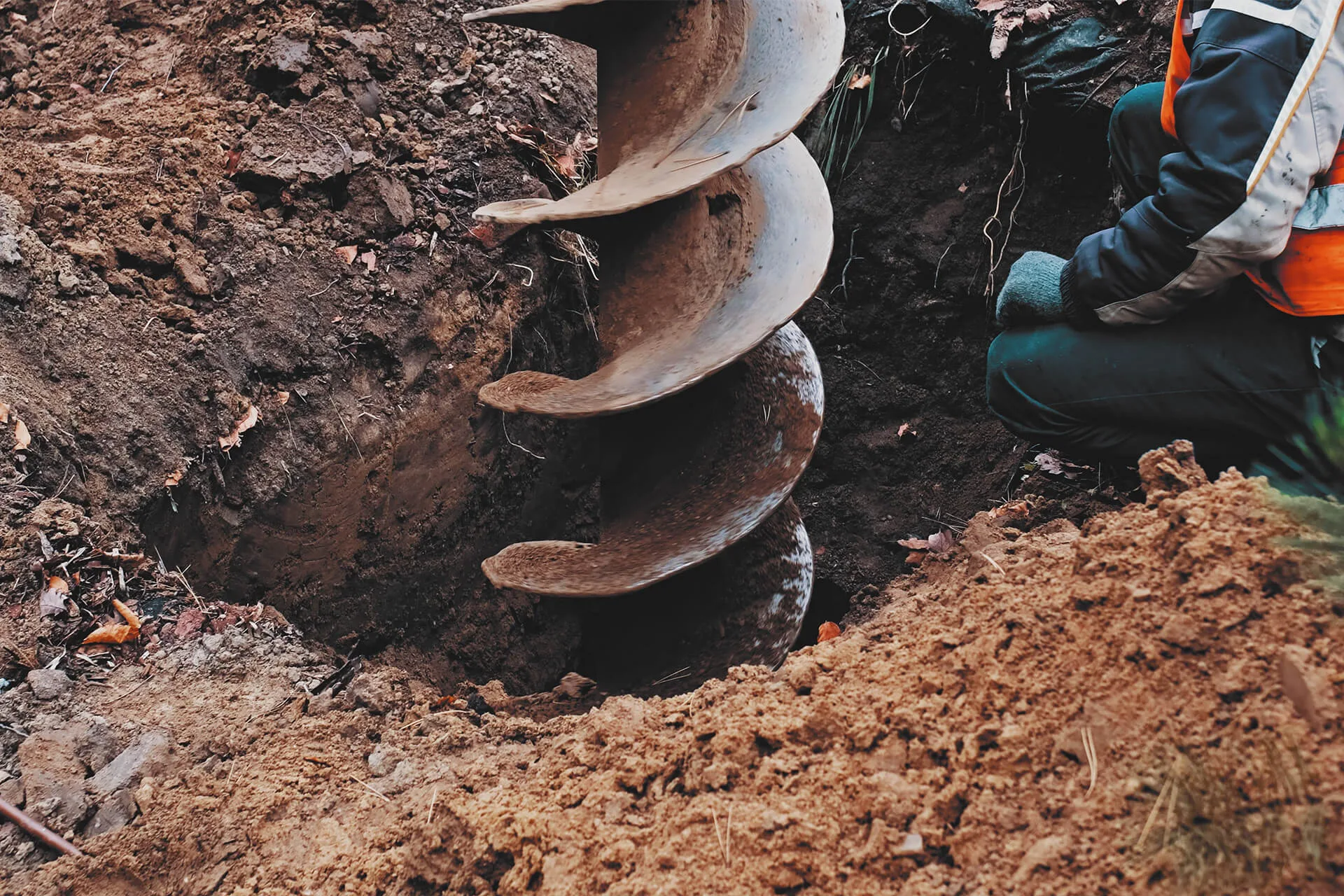 A large earth auger drills into the ground while a worker in protective gear kneels nearby, surrounded by soil and earth, as engineers conduct structural surveying of the site.