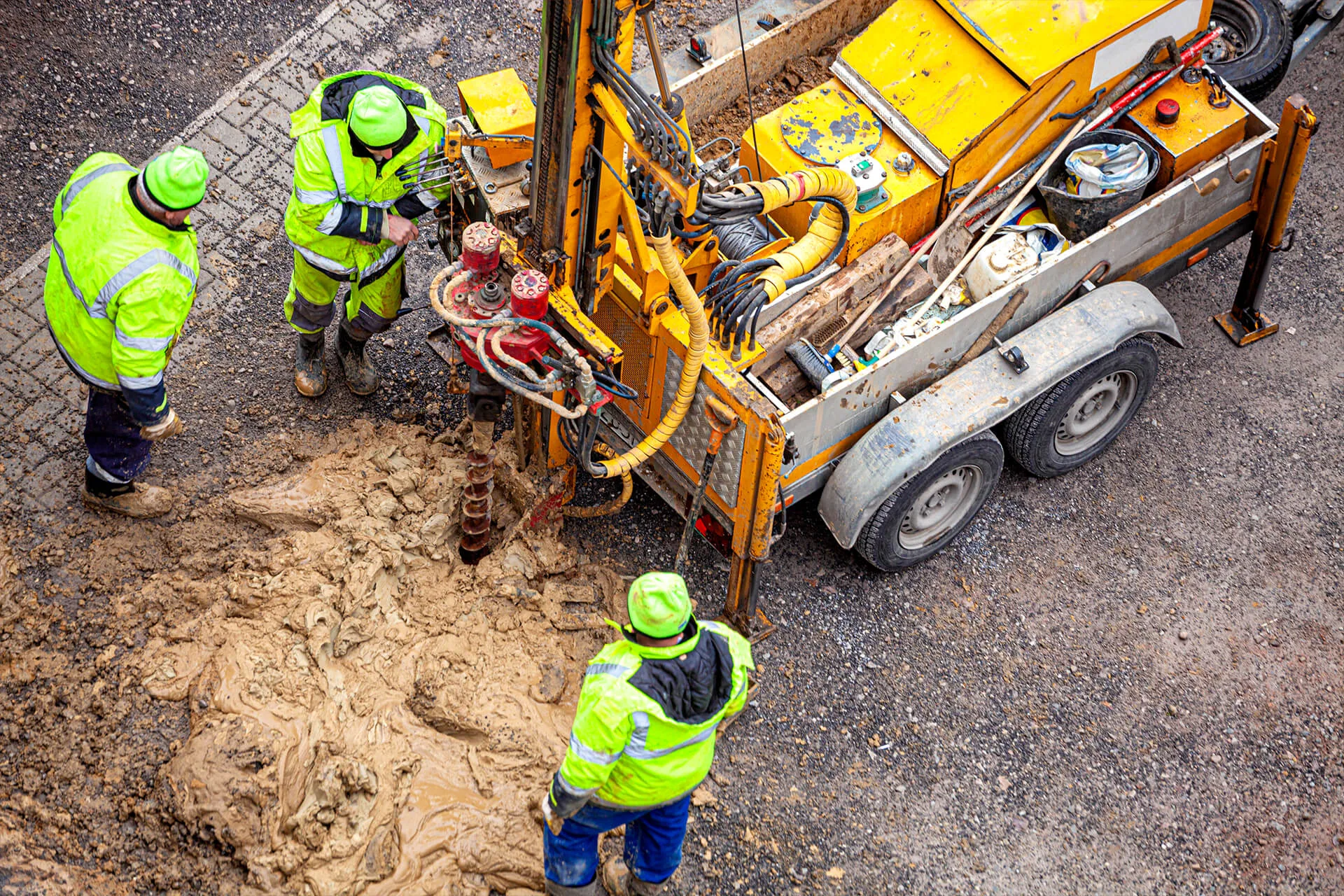 Three engineers in high-visibility clothing operate a drilling machine on a muddy construction site, surrounded by surveying equipment and wet soil.