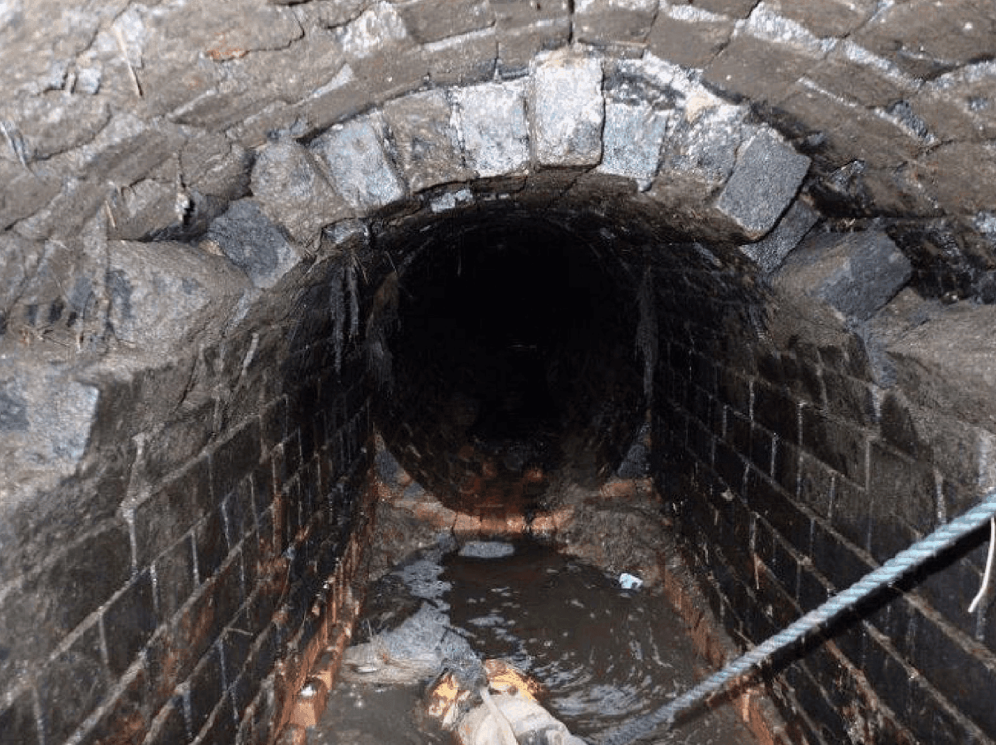 A view inside a dark, brick-lined tunnel with water and debris on the floor and a rope extending into the tunnel.