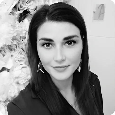 A woman with long dark hair wearing earrings and a dark top stands indoors in front of a wall decorated with large flowers.