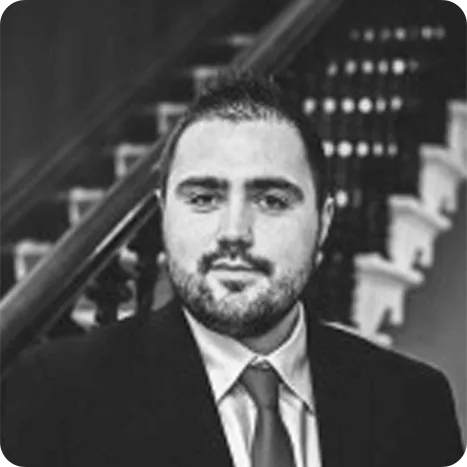 A man in a suit and tie poses in front of a staircase, looking directly at the camera. The image is in black and white.