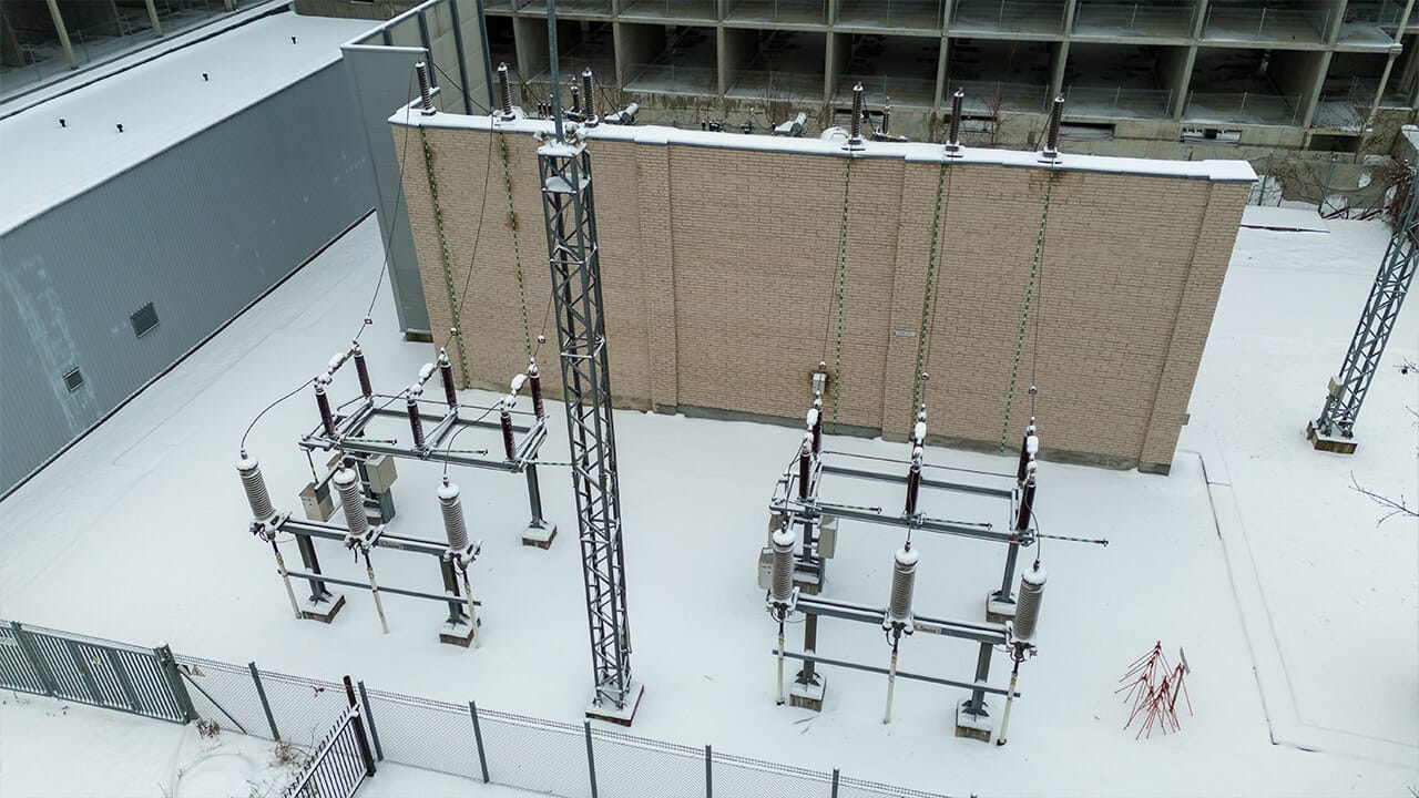 A snow-covered electrical substation with transformers and power lines next to a brick building and fenced perimeter.