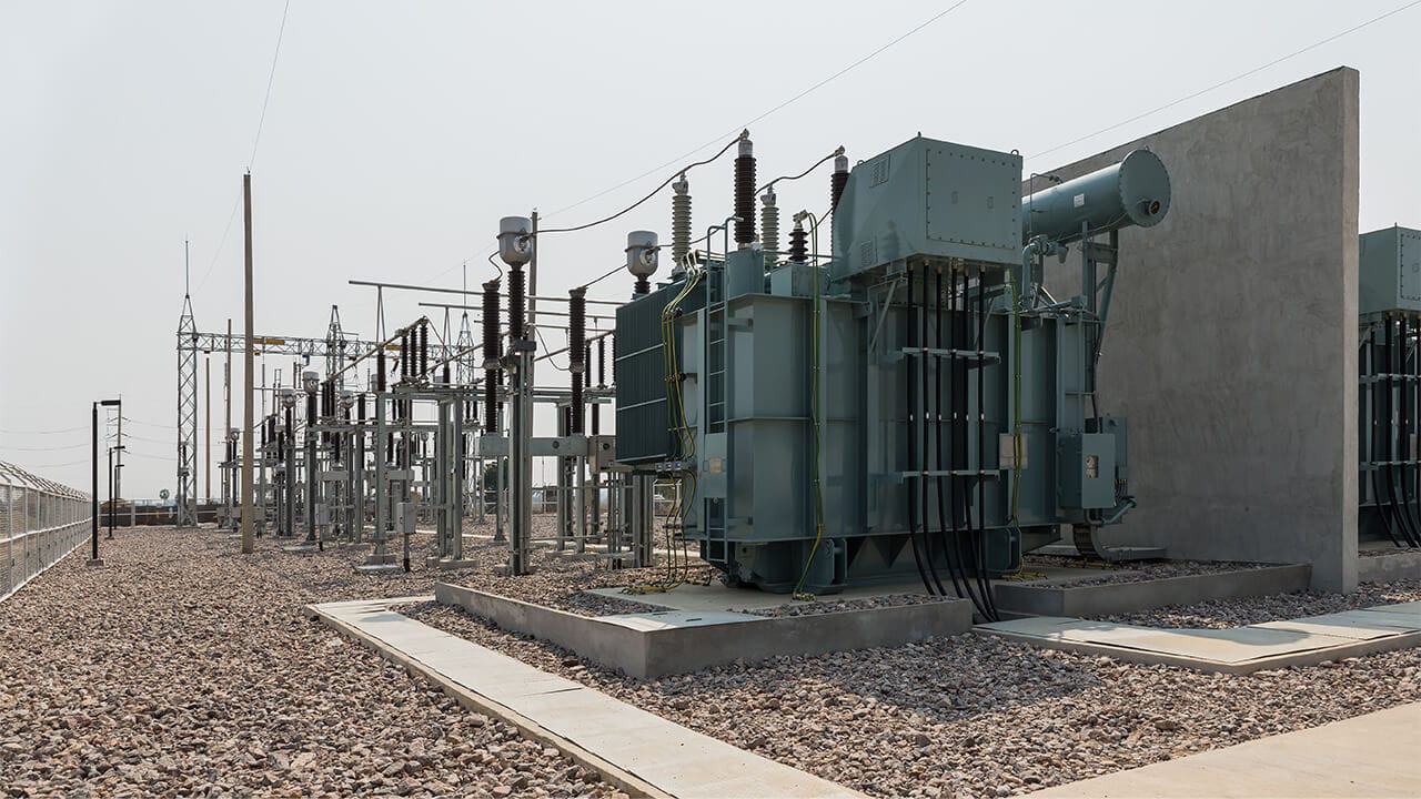 Electrical substation with large transformers and power lines, situated on a gravel car park with concrete footpaths and a clear sky overhead.