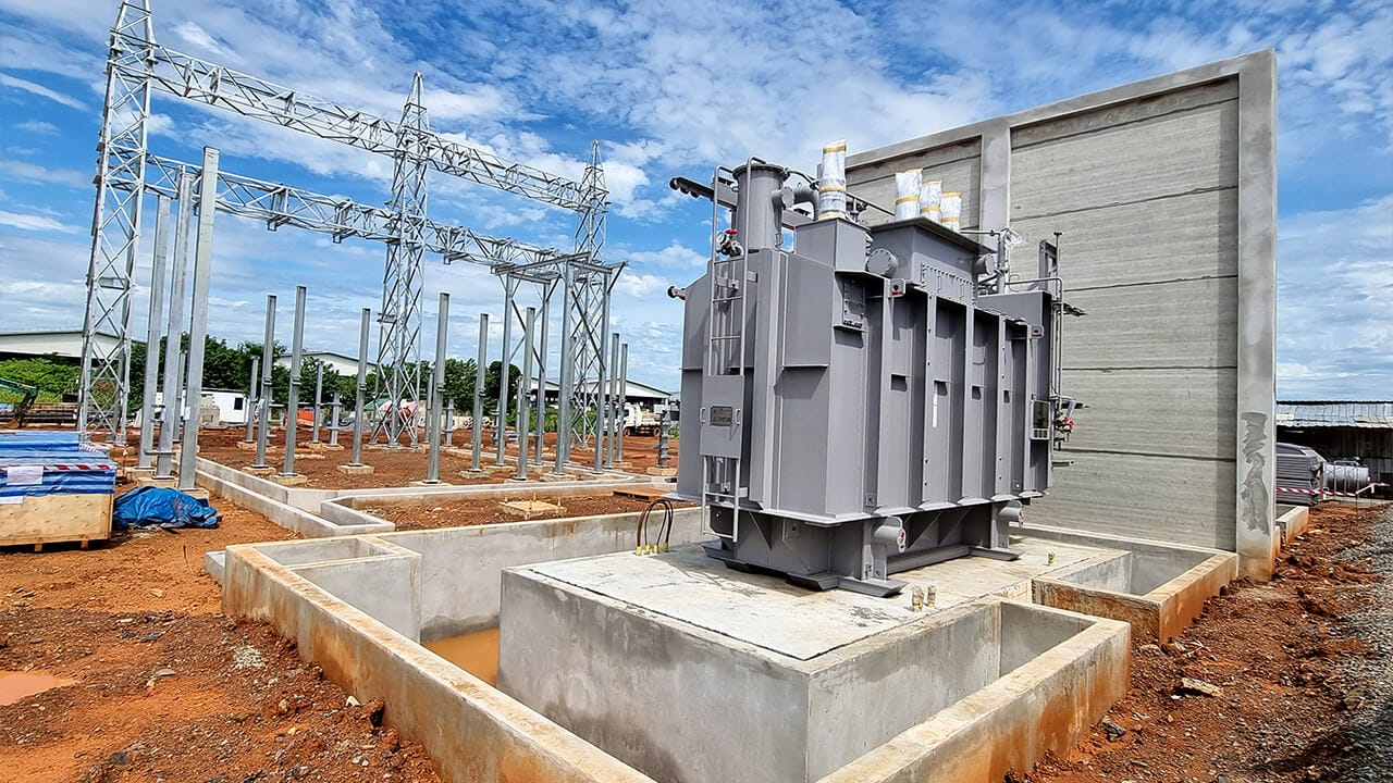 Industrial electrical substation construction site with a large power transformer mounted on a concrete foundation and metal framework in the background under a partly cloudy sky.