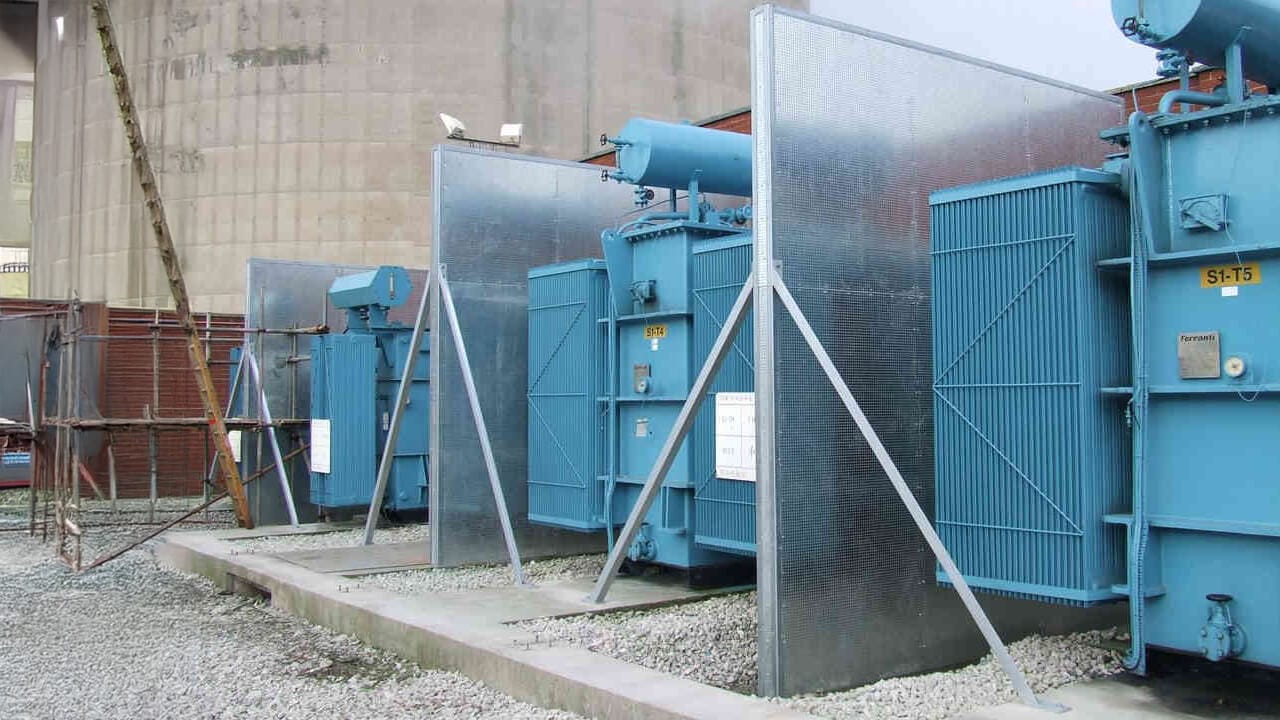 Three large blue electrical transformers are installed outdoors on concrete pads, separated by metallic fire barriers, with gravel covering the ground around them.