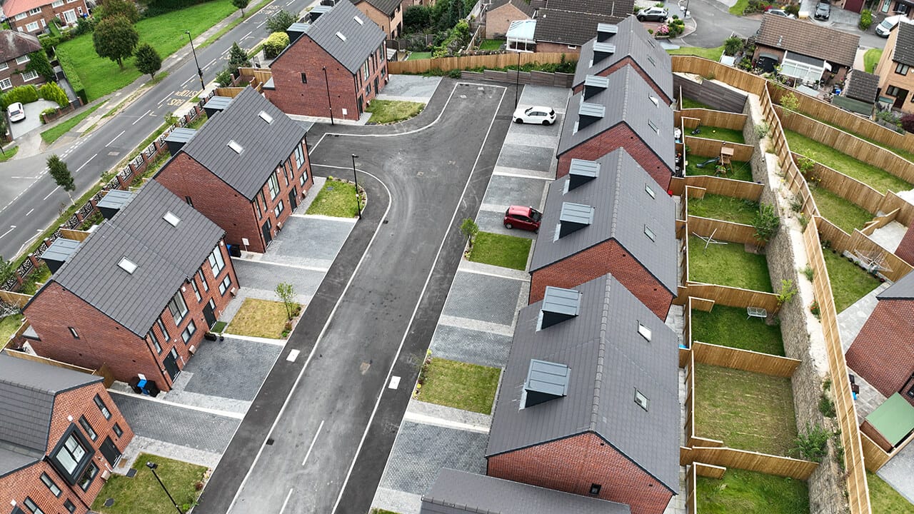 Aerial view of a modern residential neighbourhood with rows of red brick houses, driveways, cars parked, and fenced back gardens.