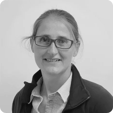 A woman with glasses and long hair tied back, wearing a collared shirt and a zip-up jacket, smiles at the camera against a plain background.