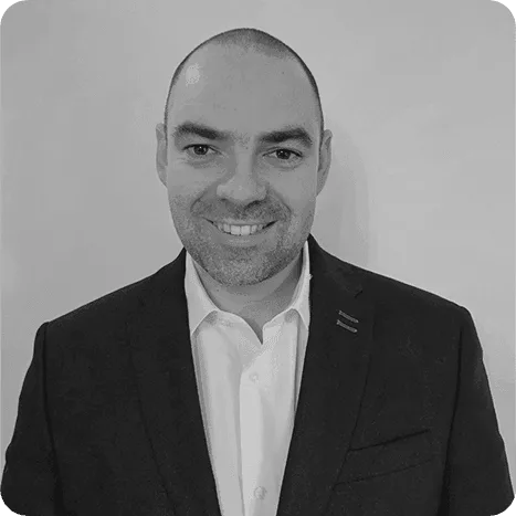 A man with a shaven head, wearing a dark suit jacket and white shirt, smiles at the camera against a plain light background.