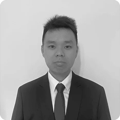 A man in a suit and tie stands against a plain, light-coloured background, looking directly at the camera.