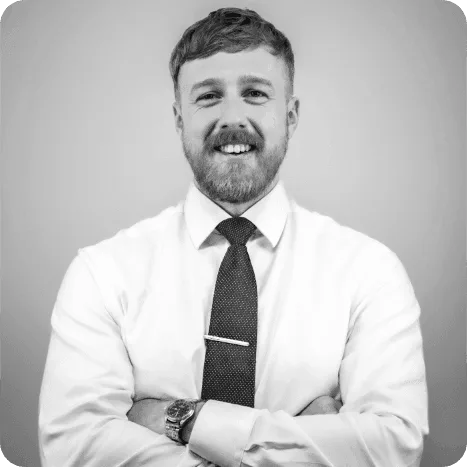 Man with a beard and moustache wearing a white shirt and dark tie, arms crossed, smiling, in front of a plain background.