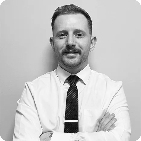 Man with short hair and moustache, wearing a white shirt and dark polka dot tie, standing with arms crossed against a plain light background.
