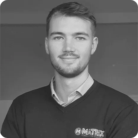 A man with short hair and a beard, wearing a collared shirt and a Matrix jumper, poses for a portrait against a plain background.