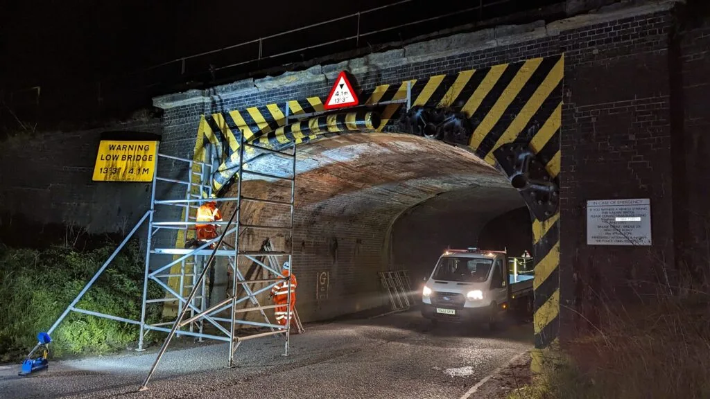 Engineers in orange PPE working from a scaffold tower at night to inspect a masonry railway underbridge marked with a 4.1 metre low bridge warning, following a bridge strike.