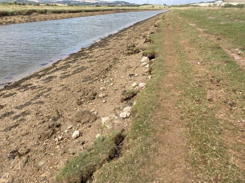 A dirt and grass path runs alongside a narrow river or canal, with rocks and erosion visible on the riverbank under a clear sky.
