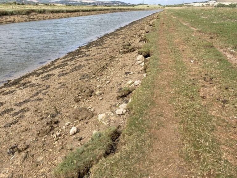 A dirt and grass path runs alongside a narrow river or canal, with rocks and erosion visible on the riverbank under a clear sky.