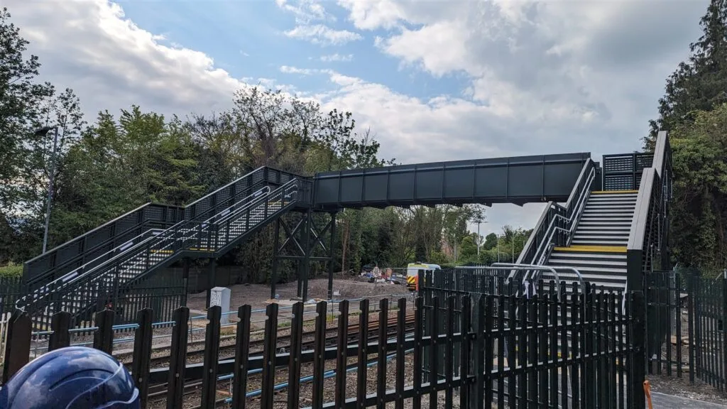A completed dark green steel footbridge spanning electrified running lines at Tottenham Hale Station, with stepped approaches on both sides and palisade fencing along the track.