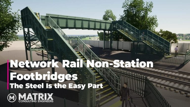A steel pedestrian footbridge spans over railway tracks, with green railings and stairs on both sides. People are walking on the bridge. Trees and a fence are in the background.