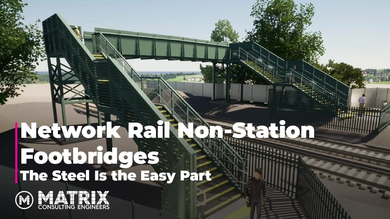 A steel pedestrian footbridge spans over railway tracks, with green railings and stairs on both sides. People are walking on the bridge. Trees and a fence are in the background.