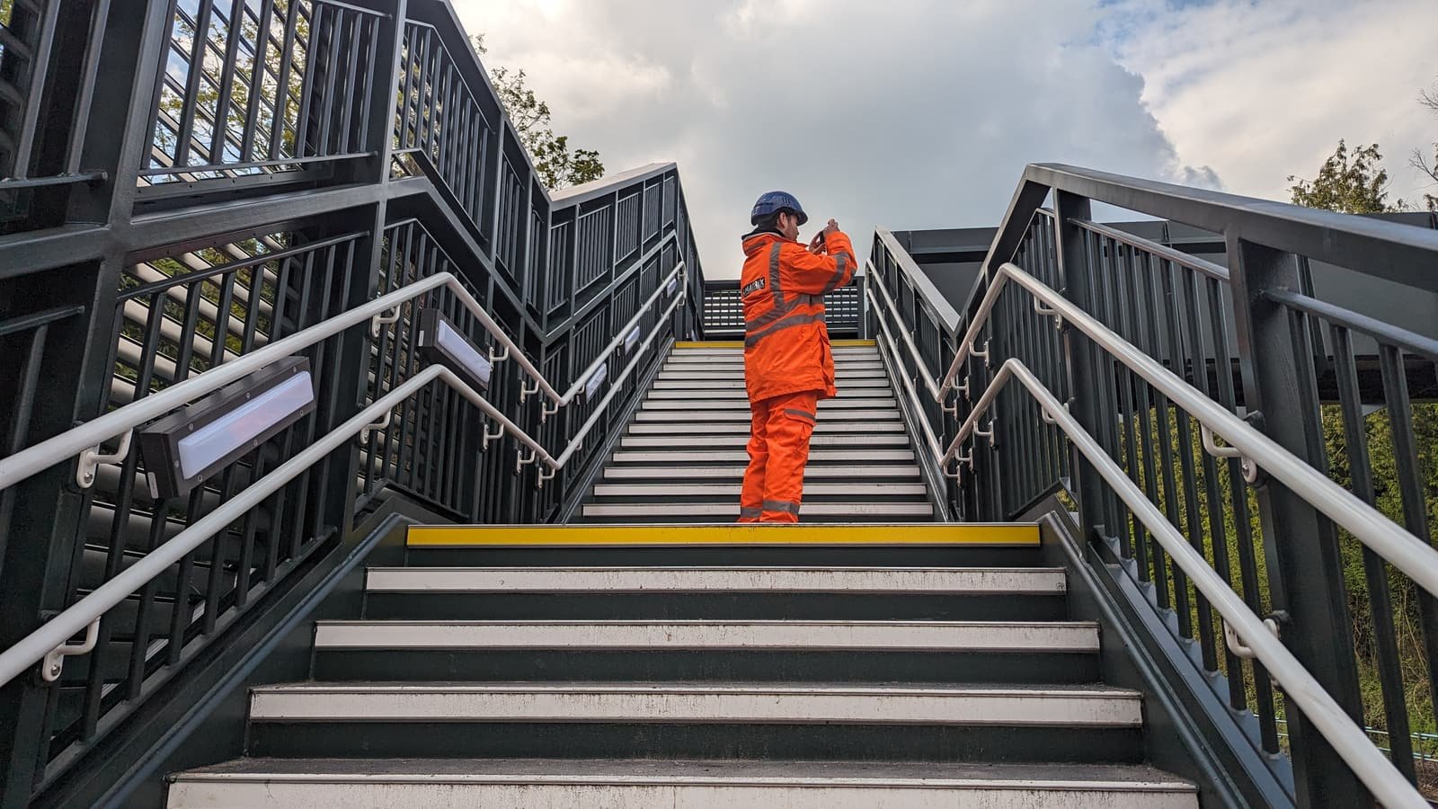 A worker in orange protective clothing descending the steps of a completed railway footbridge.