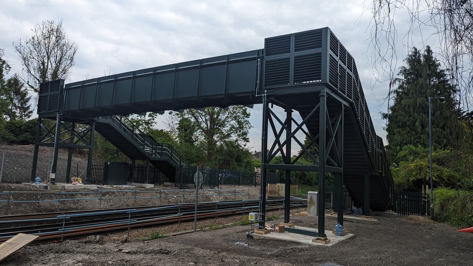 A completed dark green steel footbridge spanning two railway tracks, with stepped access on both sides.