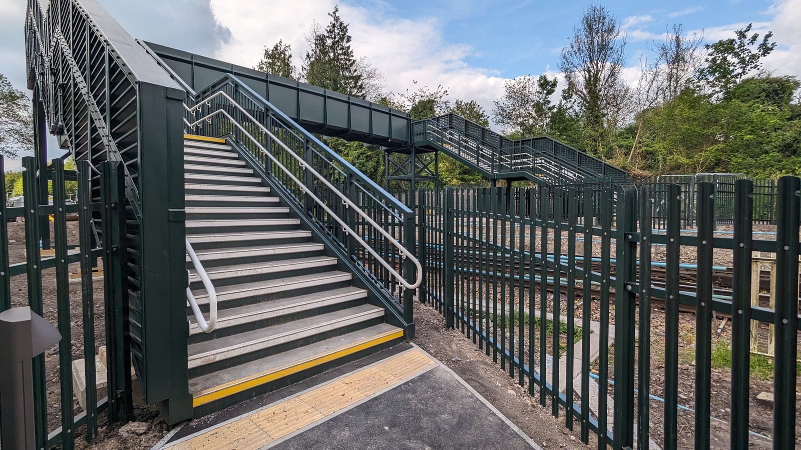 Looking up a flight of steps on a newly completed railway footbridge, with a worker in orange protective clothing on the landing above.