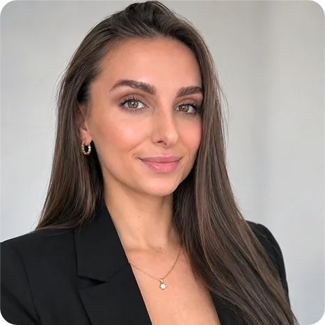 Woman with long brown hair and light eyes, wearing a black blazer and gold necklace, standing against a plain light background.