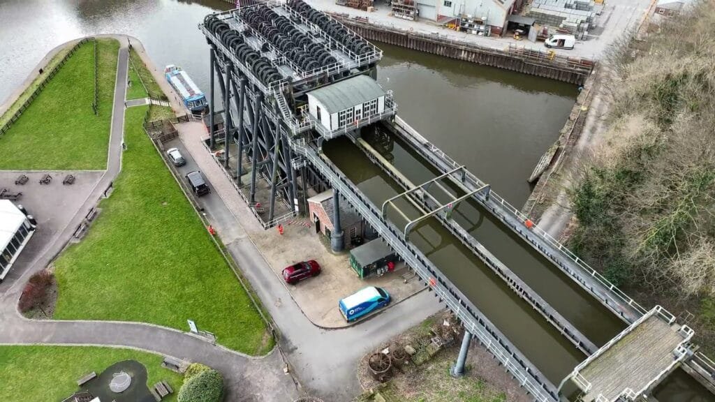 Aerial view of an industrial boat lift with water-filled chambers, adjacent to a river, grassy areas, parked cars, and nearby buildings.