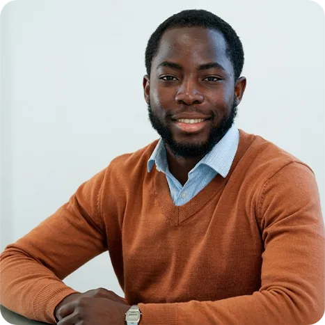 A man with a beard, wearing a light blue shirt and brown jumper, sits at a table smiling with his arms resting in front of him against a plain light background.