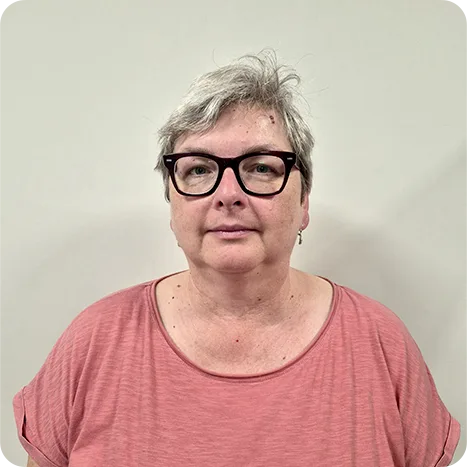 Person with short grey hair and black glasses wearing a pink top stands against a plain off-white background, looking at the camera.