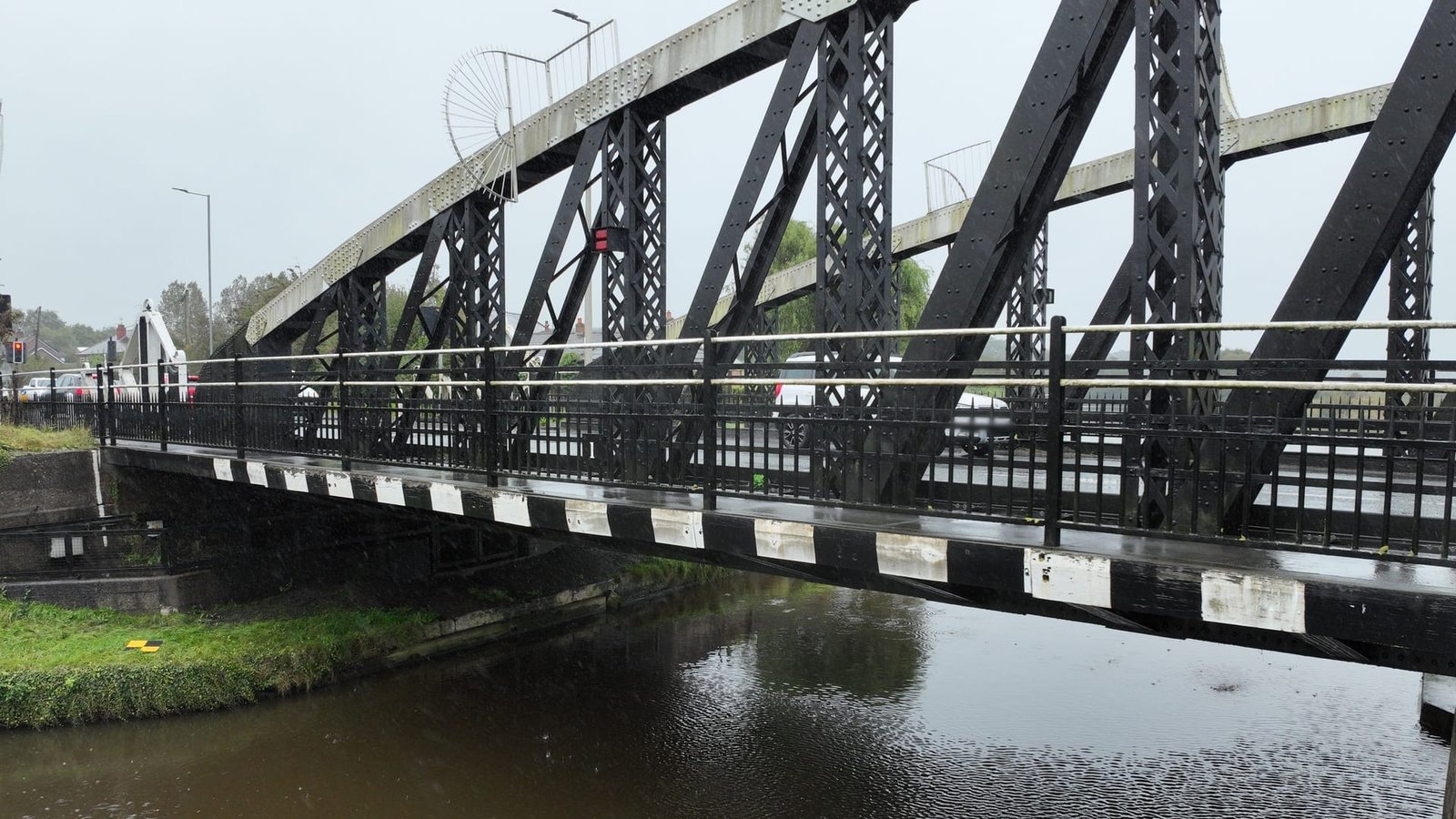 A black metal truss bridge spans over a calm river on a cloudy day, with vehicles and pedestrians visible in the background.